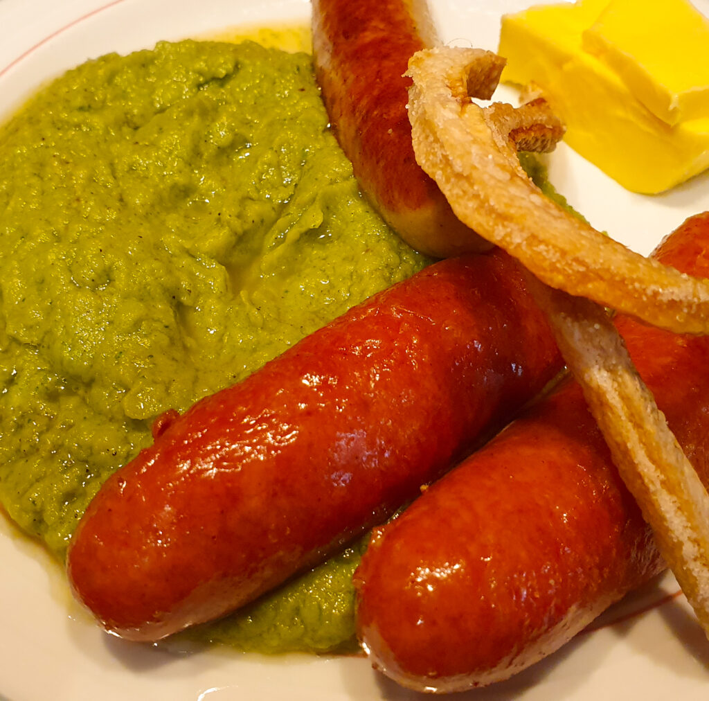 Eine Portion Brokkoli-Püree, zwei Mettenden, eine Bratwurst, Butter und zwei Schweinekrusten auf einem weißen Teller.